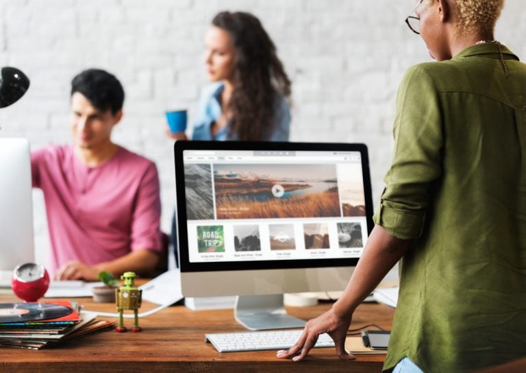 Photo of a group of people working in an office. One woman is looking at a website while two individuals in the background are working together.