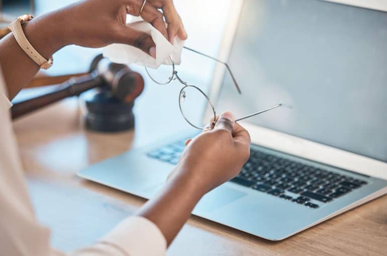 Vintage eyeglasses being cleaned with a cloth near a laptop on a wooden desk.