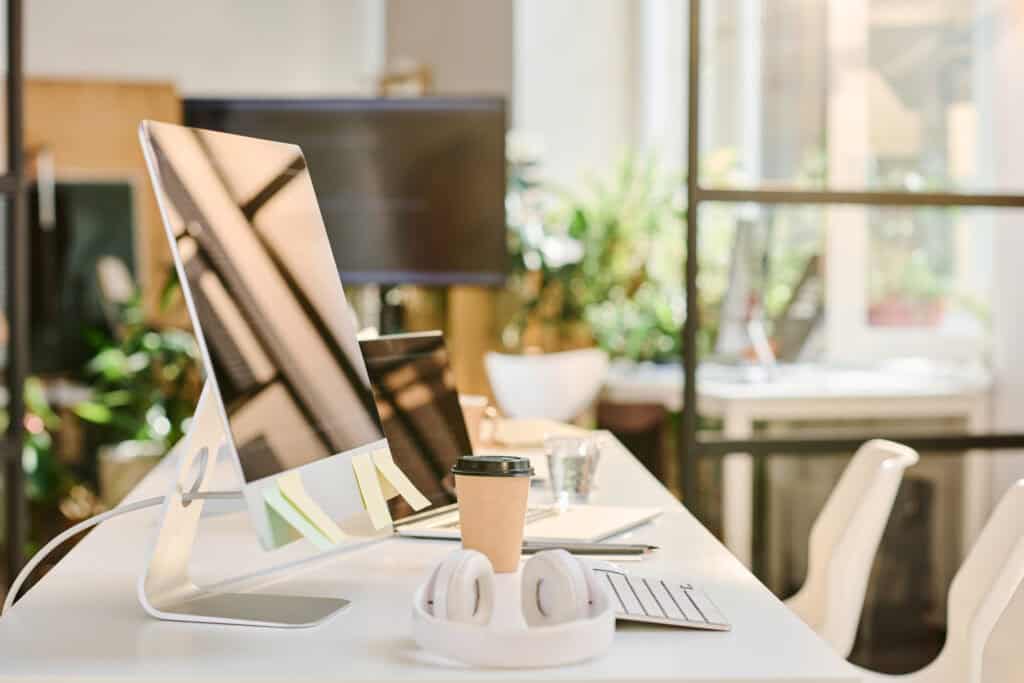 Contemporary office workspace featuring a sleek computer monitor, a keyboard, a coffee cup, and headphones on a white desk in a bright, modern office environment.
