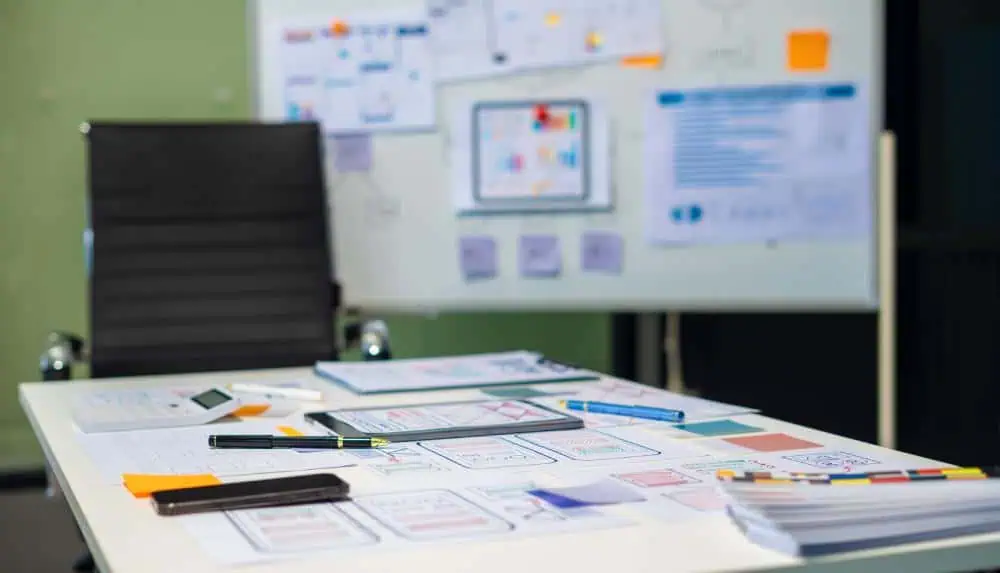Modern office workspace with design sketches, color swatches, and stationery on a white desk, with a black office chair and a whiteboard in the background.