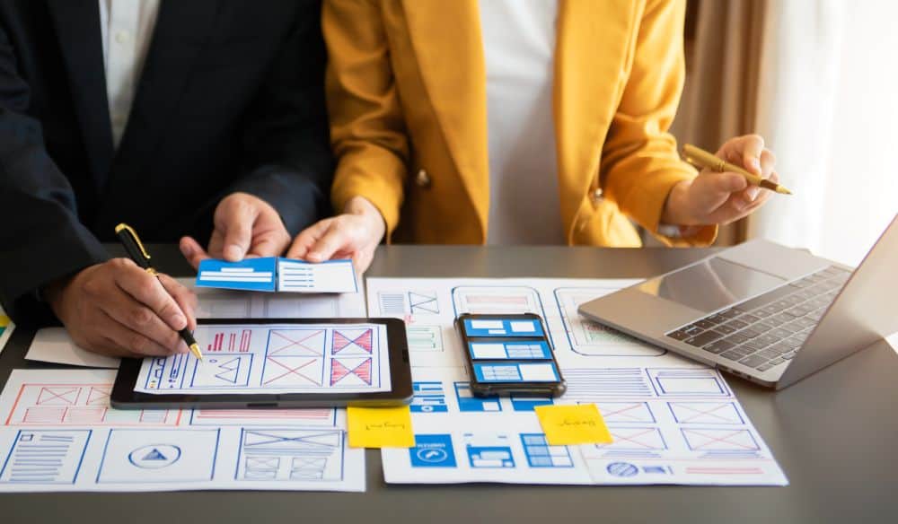 Two professionals collaborating on website design and management, reviewing wireframes, mobile app prototypes, and digital layouts on a desk.